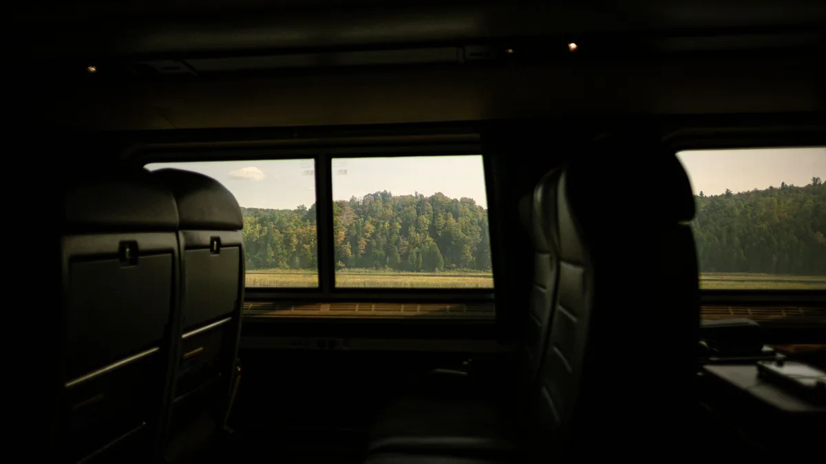 Leerer Zugwagen im Halbdunkel, Sitzlehnen von hinten, durch die Fenster eine Wiese und ein Waldrand im Tageslicht