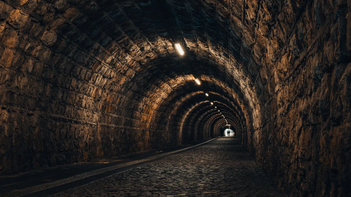 Ein gewölbter Tunnel aus Bruchstein mit Kopfsteinpflaster und Deckenlampen, ganz hinten ein heller Ausgang mit zwei kleinen Gestalten