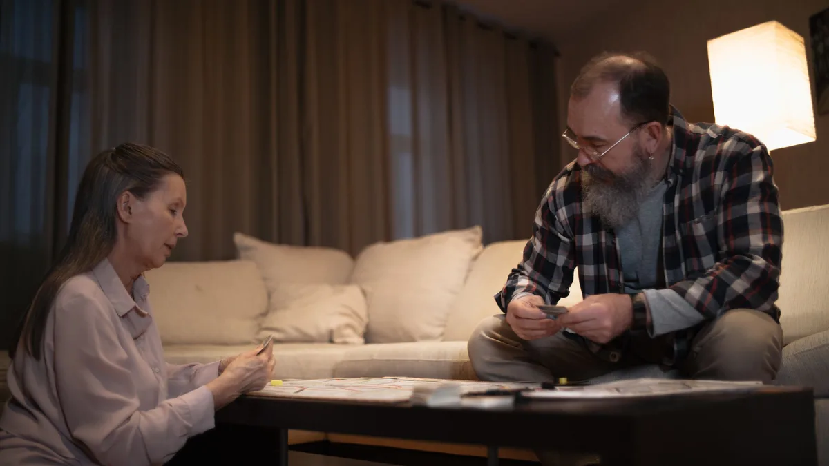 Eine Frau und ein bärtiger Mann mit Brille sitzen abends an einem niedrigen Couchtisch und halten kleine Karten in den Händen, hinter ihnen ein helles Sofa und eine Stehlampe