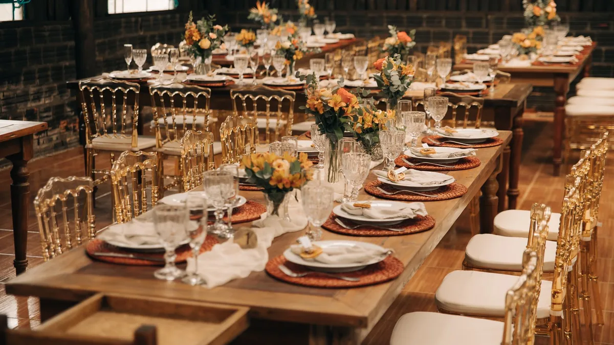 Lange gedeckte Holztafeln mit Blumengestecken, Gläsern und goldfarbenen Stühlen in einem Saal