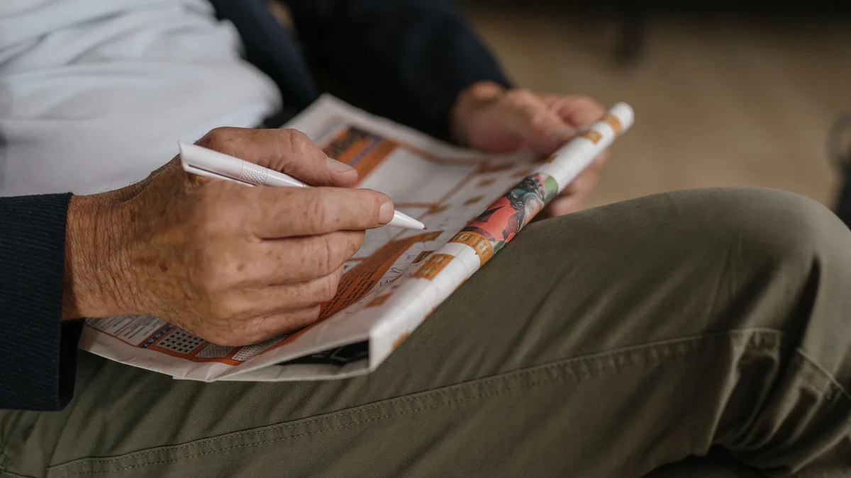 Eine ältere Hand trägt mit einem weißen Stift etwas in ein Rätsel in einer gefalteten Zeitung ein, die auf dem Oberschenkel liegt