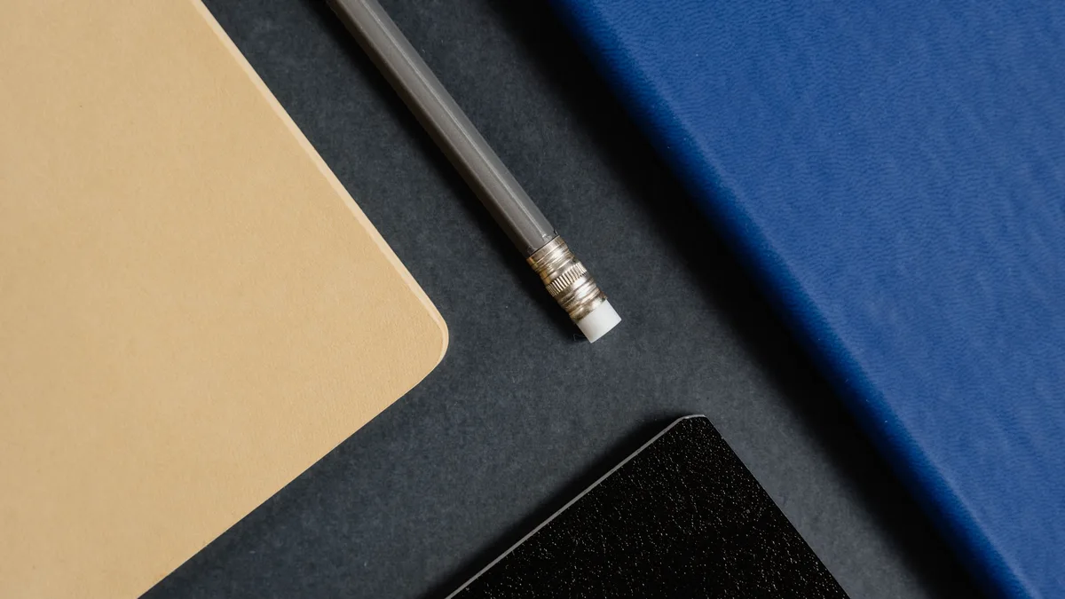 Ein Bleistift mit Radiergummi liegt zwischen einem hellbraunen und einem blauen Notizheft auf dunklem Untergrund