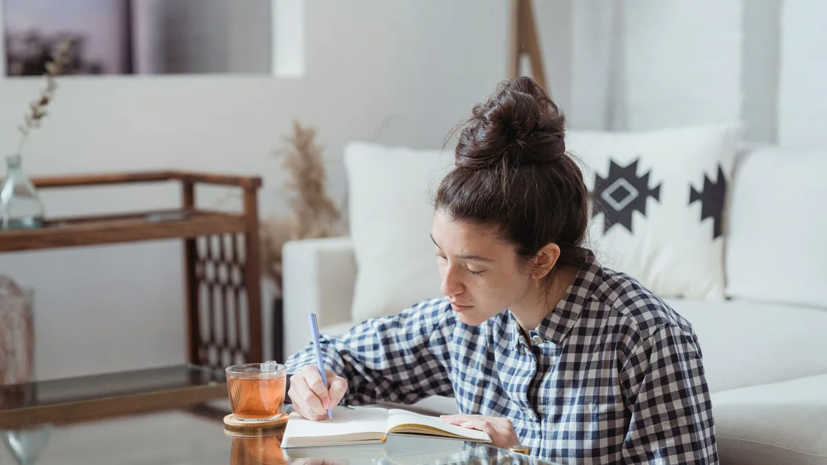 Eine junge Frau mit Haarknoten und kariertem Hemd schreibt mit einem blauen Stift in ein Notizbuch, daneben ein Glas Tee, im Hintergrund ein helles Sofa