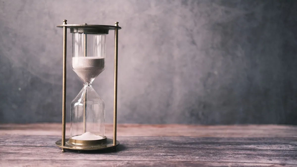 Eine Sanduhr mit Messinggestell steht auf einer verwitterten Holzplatte vor einer grauen Wand