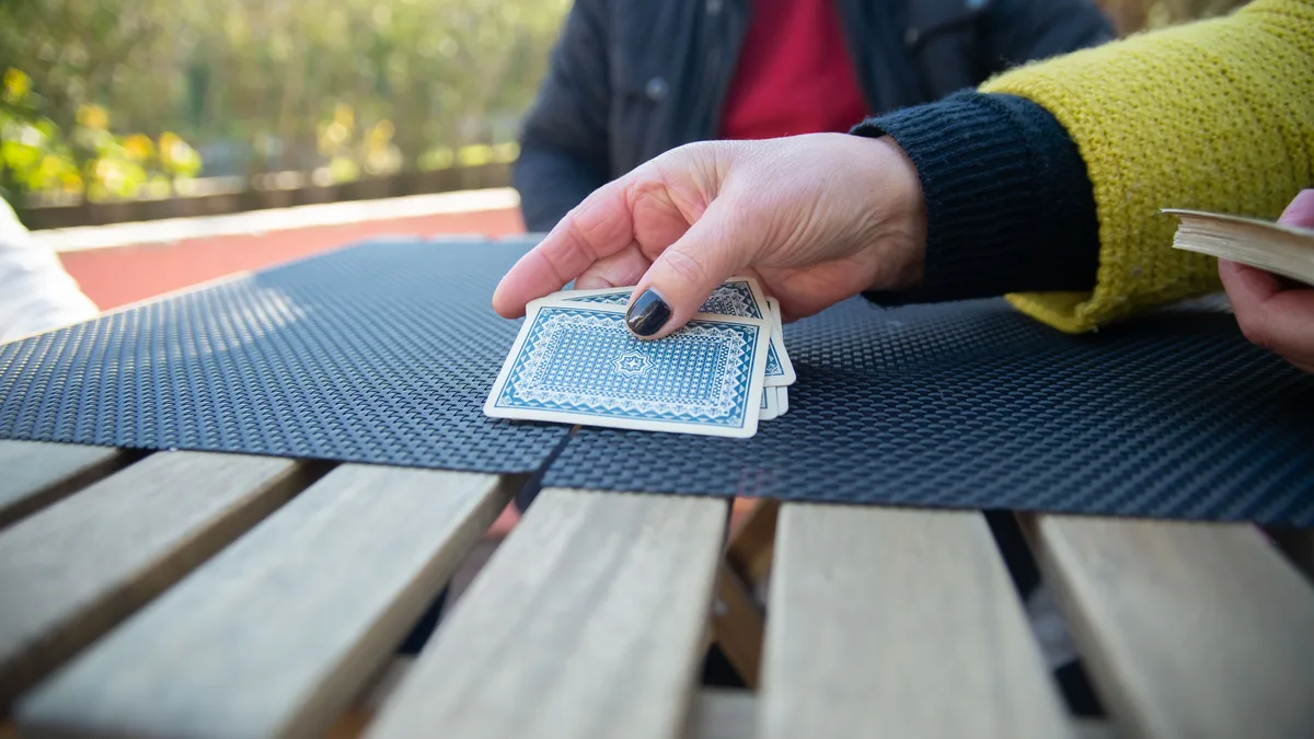 Eine Hand legt verdeckte Spielkarten mit blauem Rückenmuster auf eine dunkle Tischmatte auf einem Holztisch im Freien