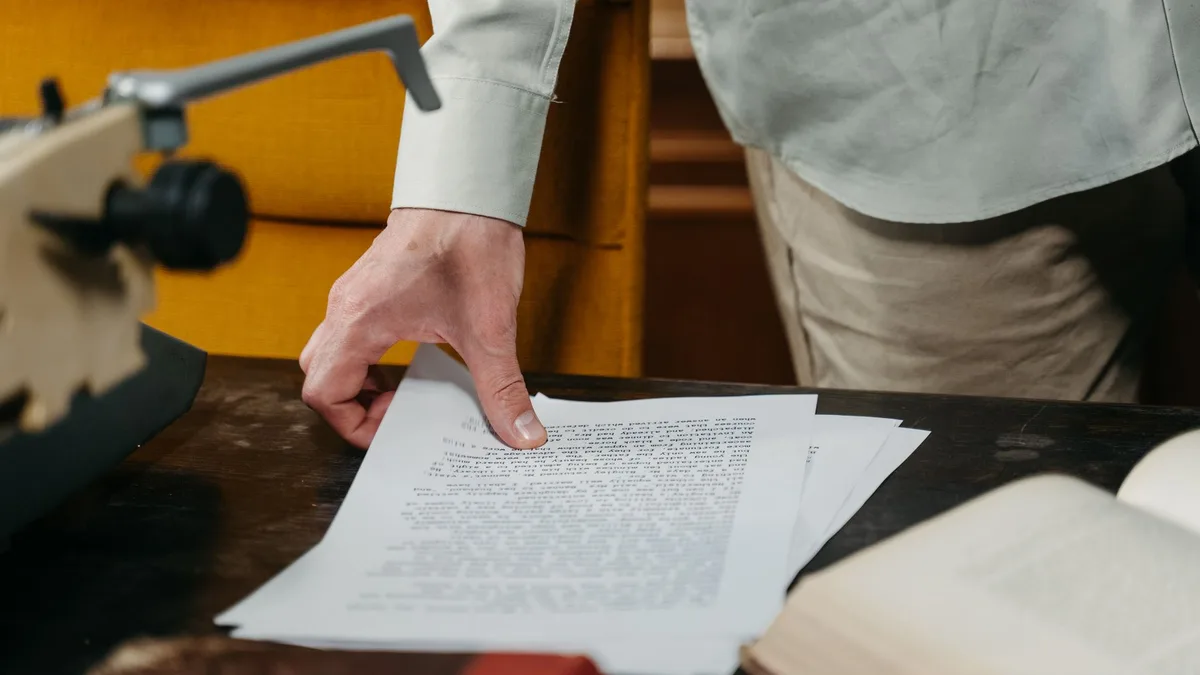 Eine Hand legt sich auf einen Stapel maschinengeschriebener Seiten auf einem dunklen Holztisch, links eine Schreibmaschine, rechts ein aufgeschlagenes Buch
