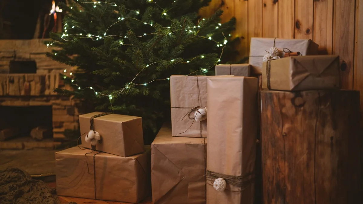 In Packpapier und Schnur verpackte Geschenke stehen unter einem Tannenbaum mit Lichterkette, daneben ein Holzklotz vor einer Holzwand