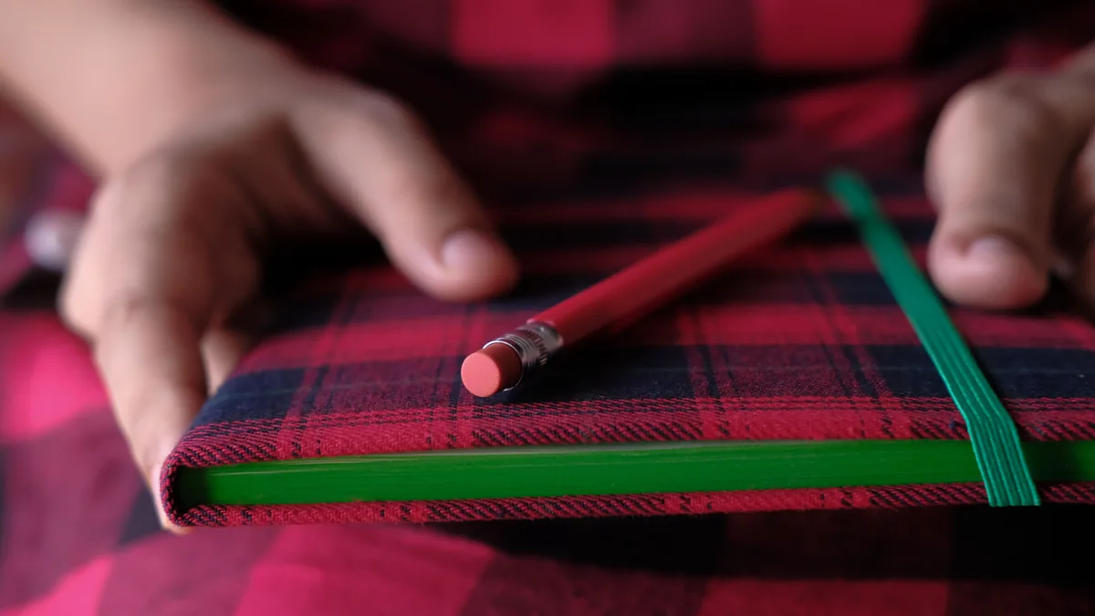 Hände halten ein rot-schwarz kariertes Notizbuch mit grünem Gummiband, darauf ein roter Bleistift