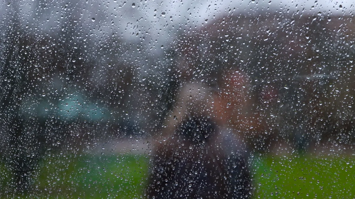 Regentropfen auf einer Fensterscheibe, dahinter verschwommen eine Person vor einer grünen Wiese